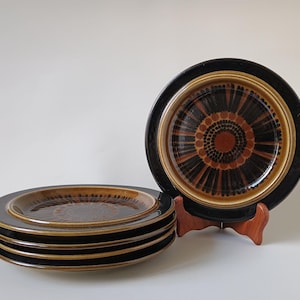 Peut inclure: Un ensemble de cinq assiettes rondes en céramique présentant une palette de couleurs brun foncé et noir. Les assiettes arborent un motif circulaire central de petits points entouré de lignes sombres rayonnantes, avec un large bord noir.