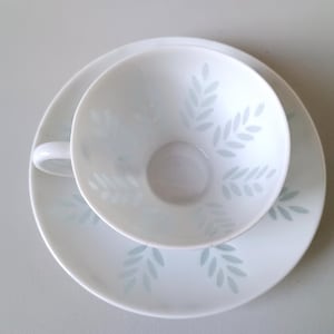 May include: A white ceramic teacup and matching saucer set featuring a delicate pattern of light blue leaf sprigs. The teacup has a rounded bowl and a small handle, while the saucer is circular with a matching leaf design around the center.