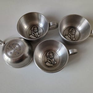Könnte beinhalten: Vier Vintage-Tassen aus silberfarbenem Metall mit Griffen. Jede Tasse zeigt ein detailliertes Relief eines bärtigen Mannes. Eine Tasse ist umgedreht und zeigt geprägten Text. Ein klassisches Set zum Servieren von Getränken.