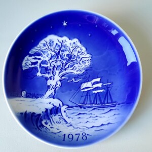 May include: A blue and white Christmas plate with a snowy tree and a ship sailing on the water. The year 1978 is printed on the bottom of the plate.