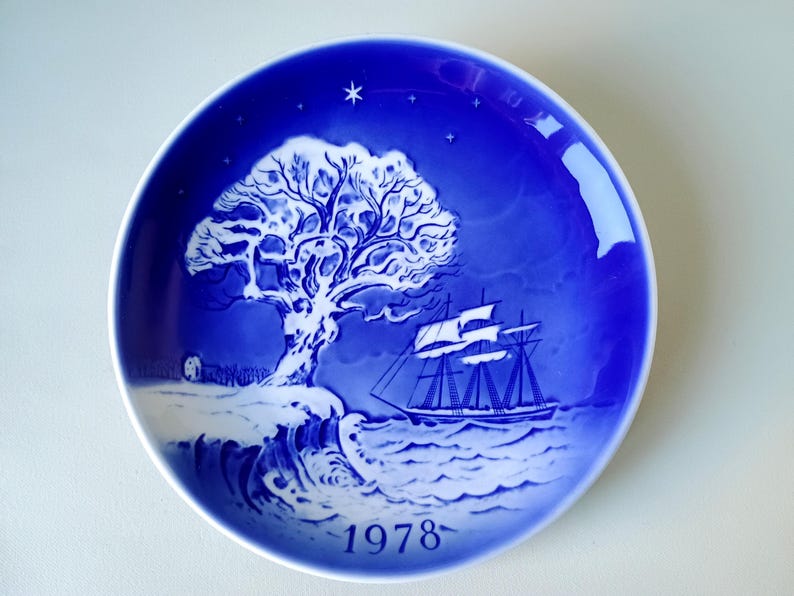 May include: A blue and white Christmas plate with a snowy tree and a ship sailing on the water. The year 1978 is printed on the bottom of the plate.