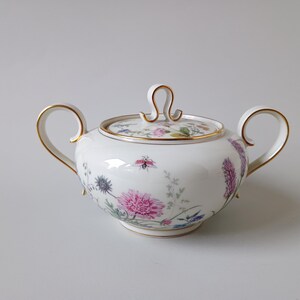 May include: White porcelain sugar bowl with lid, featuring a floral design in pink, purple, and blue. The bowl has two handles and gold trim. The lid has a loop handle and gold trim. A decorative piece for serving sugar.