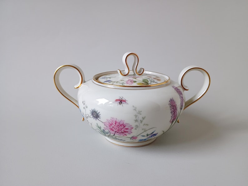 May include: White porcelain sugar bowl with lid, featuring a floral design in pink, purple, and blue. The bowl has two handles and gold trim. The lid has a loop handle and gold trim. A decorative piece for serving sugar.