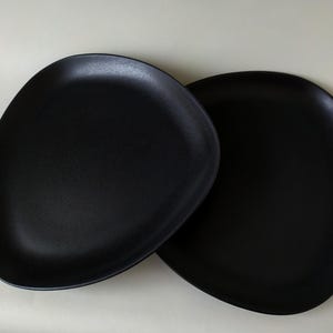 Könnte beinhalten: Zwei mattschwarze Keramikplatten mit einer organischen, asymmetrischen Form. Die Platten haben eine glatte, leicht strukturierte Oberfläche und eine tiefe, solide schwarze Farbe. Die Platten sind zum Servieren von Speisen bestimmt.