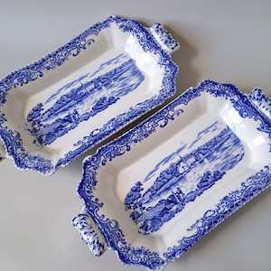 May include: Two rectangular white ceramic serving trays featuring a blue transferware pattern. Each tray has ornate blue scrollwork borders and a central landscape illustration depicting a river scene with buildings, trees, and figures.