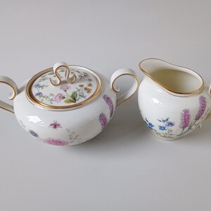 May include: A white porcelain sugar bowl with a lid and a creamer. Both pieces are decorated with floral patterns in pink, purple, and blue, and have gold trim. The sugar bowl has a lid with a gold handle.