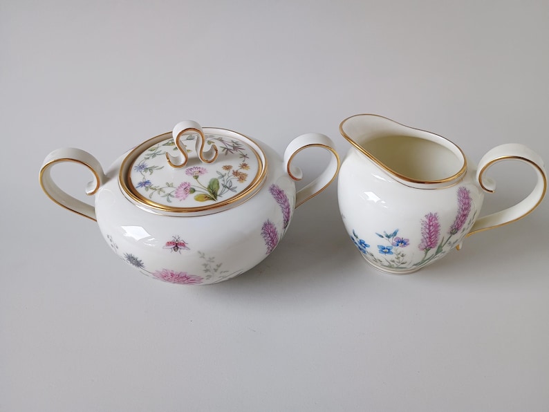 May include: A white porcelain sugar bowl with a lid and a creamer. Both pieces are decorated with floral patterns in pink, purple, and blue, and have gold trim. The sugar bowl has a lid with a gold handle.