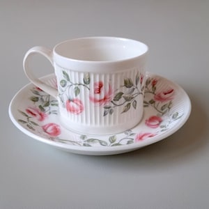 May include: A white teacup and saucer with a floral pattern of pink roses and green leaves. The cup and saucer have a ribbed design.