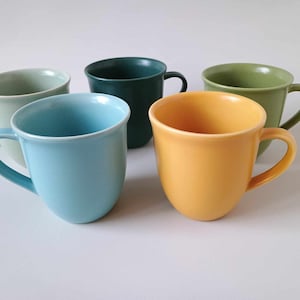 Op de afbeelding: Een set van vijf keramische mokken in verschillende effen kleuren, waaronder lichtblauw, felgeel, donker bosgroen, saliegroen en licht mintgroen. Elke mok heeft een glad, afgerond lichaam met een gebogen handvat en een eenvoudig, strak ontwerp.
