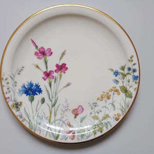 May include: A white ceramic plate with a gold rim, decorated with a floral design. The flowers include pink, blue, and yellow blooms, with green foliage. The plate is round and has a smooth surface.