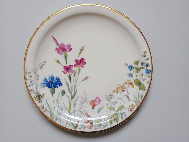May include: A white ceramic plate with a gold rim, decorated with a floral design. The flowers include pink, blue, and yellow blooms, with green foliage. The plate is round and has a smooth surface.