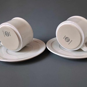 K&ouml;nnte beinhalten: Zwei wei&szlig;e Teetassen und Untertassen aus Keramik mit einem dezenten grauen Streifen am Rand der Untertassen. Die Unterseite jeder Tasse tr&auml;gt ein kreisf&ouml;rmiges Logo mit dem Text "Royal Copenhagen" und einer Krone.