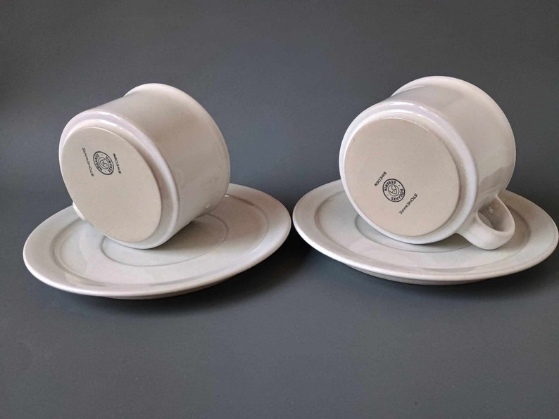 K&ouml;nnte beinhalten: Zwei wei&szlig;e Teetassen und Untertassen aus Keramik mit einem dezenten grauen Streifen am Rand der Untertassen. Die Unterseite jeder Tasse tr&auml;gt ein kreisf&ouml;rmiges Logo mit dem Text "Royal Copenhagen" und einer Krone.