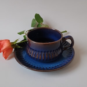 May include: A blue ceramic teacup and saucer set with a brown rim. The cup has a ribbed design and a handle. A coral rose and green leaves are next to the cup and saucer. The set is on a white surface.