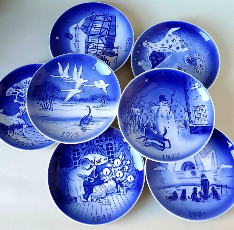 May include: A collection of seven blue and white porcelain Christmas plates featuring various holiday scenes. The plates are decorated with white outlines and have the year of production printed on the bottom. The plates include scenes of a house, a snowman, a swan, a Christmas tree, and a winter landscape.