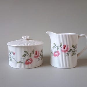 Könnte beinhalten: Ein weißes Zucker- und Milchkännchen-Set aus Keramik mit einem Blumendesign. Das Set zeigt rosa Rosen und grüne Blätter auf weißem Hintergrund. Die Zuckerdose hat einen Deckel mit einem dekorativen Knopf.