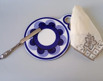 May include: A round, white and blue floral-patterned serving plate with a handle, a silver butter knife, and a folded white linen napkin with embroidered floral details. The plate has a dark blue border and a central white circle.