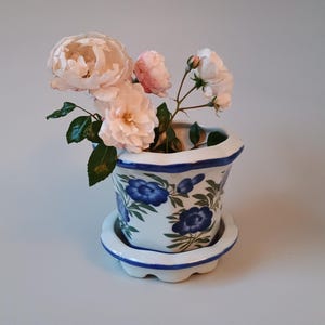 Pode incluir: Um vaso de flores de cerâmica branca com borda recortada e pires a condizer, decorado com padrões florais azuis pintados à mão e folhas verdes. O vaso contém um pequeno ramo de rosas cor-de-rosa claro com caules e folhagem verdes.