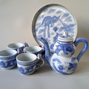 Può includere: Un set da tè in porcellana blu e bianca composto da una teiera a forma di zucca con coperchio, tre tazze abbinate con manici a forma di drago e un vassoio rotondo. Ogni pezzo è decorato con motivi di draghi e nuvole dipinti in blu.