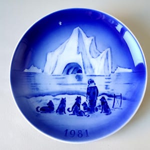 May include: A blue and white Royal Copenhagen Christmas plate featuring a snowy landscape with an iceberg and a group of people and dogs. The plate is dated 1981.