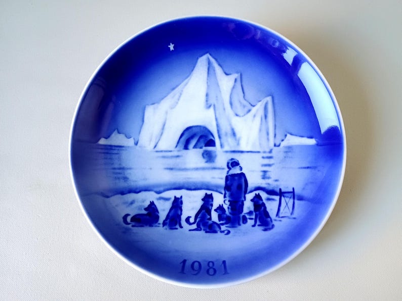 May include: A blue and white Royal Copenhagen Christmas plate featuring a snowy landscape with an iceberg and a group of people and dogs. The plate is dated 1981.