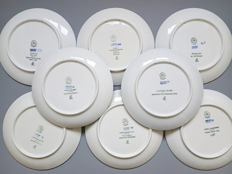 Puede incluir: Una colecci&oacute;n de platos de cer&aacute;mica blanca con sellos decorativos azules y verdes y texto. Los platos presentan varios dise&ntilde;os y textos, incluyendo "Christmas Rose and Cat" y "Winter Wonderland". Los platos est&aacute;n dispuestos sobre una superficie gris.