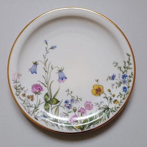May include: White ceramic plate with a gold rim, decorated with a floral design. The flowers include bluebells, clover, and other wildflowers in shades of pink, yellow, and blue. The plate has a delicate, elegant appearance.