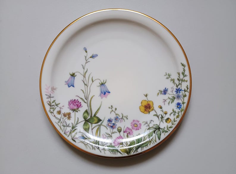 May include: White ceramic plate with a gold rim, decorated with a floral design. The flowers include bluebells, clover, and other wildflowers in shades of pink, yellow, and blue. The plate has a delicate, elegant appearance.