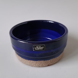 May include: A round, ceramic bowl with a deep blue interior and exterior. The bowl has a textured, light brown base and a dark blue band around the middle. A small, rectangular label with text is affixed to the side. The bowl is likely for decorative or functional use.