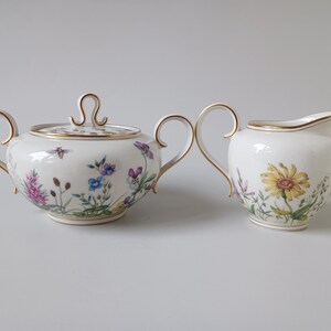May include: A white porcelain sugar bowl with a lid and a creamer. Both pieces are decorated with floral and insect motifs, and have gold trim. The sugar bowl has a bee on the side. The creamer has a yellow flower.