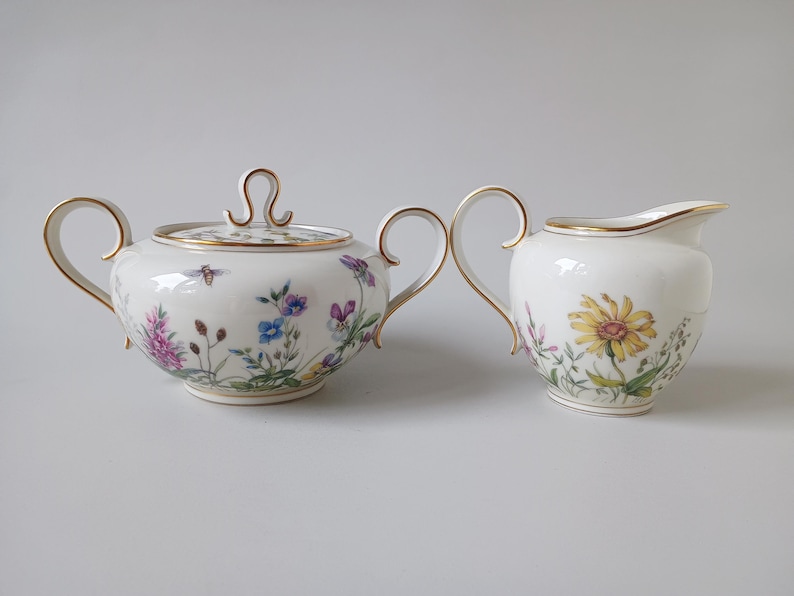 May include: A white porcelain sugar bowl with a lid and a creamer. Both pieces are decorated with floral and insect motifs, and have gold trim. The sugar bowl has a bee on the side. The creamer has a yellow flower.