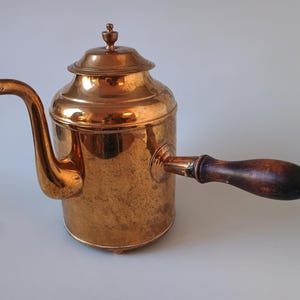 Könnte beinhalten: Eine Kaffeekanne aus poliertem Kupfer mit zylindrischem Körper, gebogenem Ausguss und abnehmbarem Deckel mit kleinem Knauf. Ein dunkelbrauner, gedrehter Holzgriff ist an der Seite befestigt und bildet einen Kontrast zur metallischen Oberfläche.