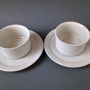 K&ouml;nnte beinhalten: Zwei wei&szlig;e Teetassen aus Keramik mit passenden Untertassen. Die Tassen haben ein schlichtes, abgerundetes Design und stehen auf einer grauen Oberfl&auml;che.
