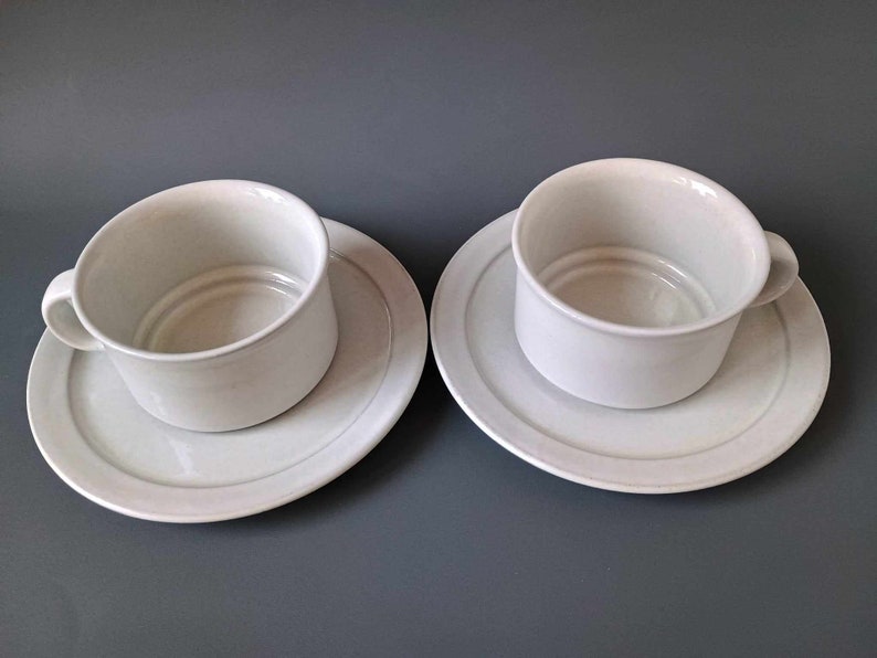 K&ouml;nnte beinhalten: Zwei wei&szlig;e Teetassen aus Keramik mit passenden Untertassen. Die Tassen haben ein schlichtes, abgerundetes Design und stehen auf einer grauen Oberfl&auml;che.