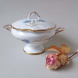 May include: A white porcelain tureen with a pedestal base, two side handles, and a matching lid. The piece features delicate blue cornflower motifs and gold trim along the scalloped edges, handles, lid finial, and the circular base.