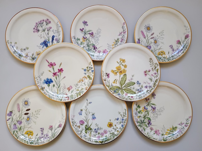 May include: A collection of eight white ceramic plates with gold rims. Each plate features a different floral design in shades of purple, pink, yellow, and blue. The plates are arranged in a circular pattern.