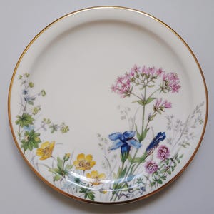May include: A white ceramic plate with a gold rim, decorated with a floral design. The flowers include yellow, blue, and pink blooms with green foliage. The plate is round and has a smooth surface.