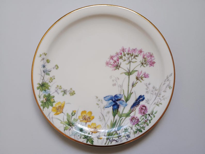 May include: A white ceramic plate with a gold rim, decorated with a floral design. The flowers include yellow, blue, and pink blooms with green foliage. The plate is round and has a smooth surface.
