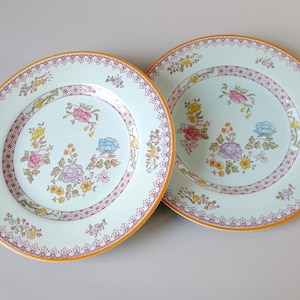 May include: Two round ceramic plates with a pale mint green base color. Each plate features a central floral arrangement in pink, purple, blue, and yellow, surrounded by a decorative maroon lattice border and a solid orange rim edge.