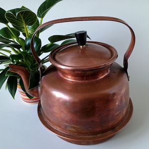 Puede incluir: Una tetera de cobre vintage con un pomo negro en la tapa. La tetera tiene un cuerpo redondeado y un pico en el lateral. La tetera está sobre una superficie blanca.