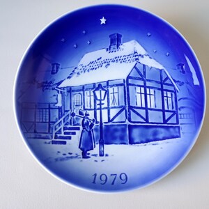 May include: A blue and white Christmas plate with a snowy house and a person holding a lantern. The plate has a blue background with white stars and the year 1979 printed on the bottom.