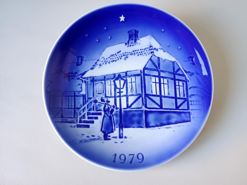 May include: A blue and white Christmas plate with a snowy house and a person holding a lantern. The plate has a blue background with white stars and the year 1979 printed on the bottom.