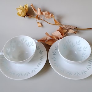 Peut inclure: Deux tasses à thé en porcelaine blanche avec soucoupes assorties ornées d'un motif délicat de feuilles gris clair. Les tasses ont une forme arrondie avec une petite anse sur le côté. Une tige de rose jaune séchée et fanée est posée dessus.