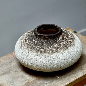 Peut inclure: Un vase en céramique texturé, avec un corps large et arrondi et un bord marron foncé. Le vase a une base blanche qui s'estompe en marron moucheté vers le haut. Il repose sur une surface en bois, mettant en valeur son design unique.