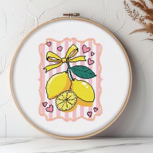 May include: A cross-stitch embroidery hoop featuring a lemon design. The design includes yellow lemons, a yellow bow, green leaves, and pink hearts on a white background with pink stripes. The embroidery is set in a wooden hoop.