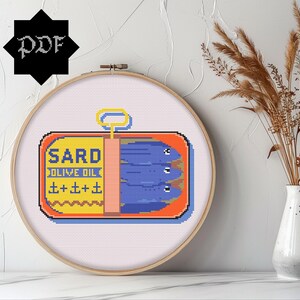 May include: A cross-stitch pattern of a sardine can with the text "SARD OLIVE OIL" and a blue and white illustration of sardines inside the can.