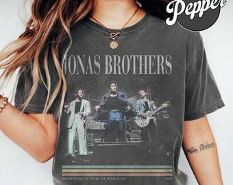Op de afbeelding: Een antracietgrijs T-shirt met korte mouwen en een vintage-stijl afbeelding van de Jonas Brothers op het podium. De tekst luidt JONAS BROTHERS, THE BURNING UP TOUR: ALL OVER AGAIN en 2026, met een meerkleurig horizontaal streepontwerp.