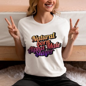 Op de afbeelding: Wit T-shirt met de tekst "Natural born Alpha Male Slayer" in een verloop van oranje, rood en paars. Het shirt is een casual stijl, geschikt voor dagelijks gebruik.