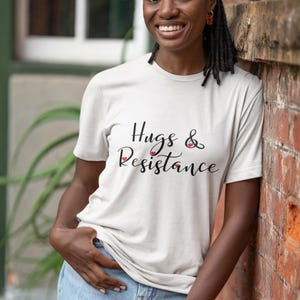 May include: A light-colored t-shirt with the words "Hugs & Resistance" in a script font. The shirt is worn with light blue jeans. The person is leaning against a brick wall.