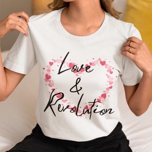 May include: White t-shirt with the words "Love & Revolution" in black script. The word "Love" is above the ampersand, and "Revolution" is below. A heart made of pink hearts surrounds the words.